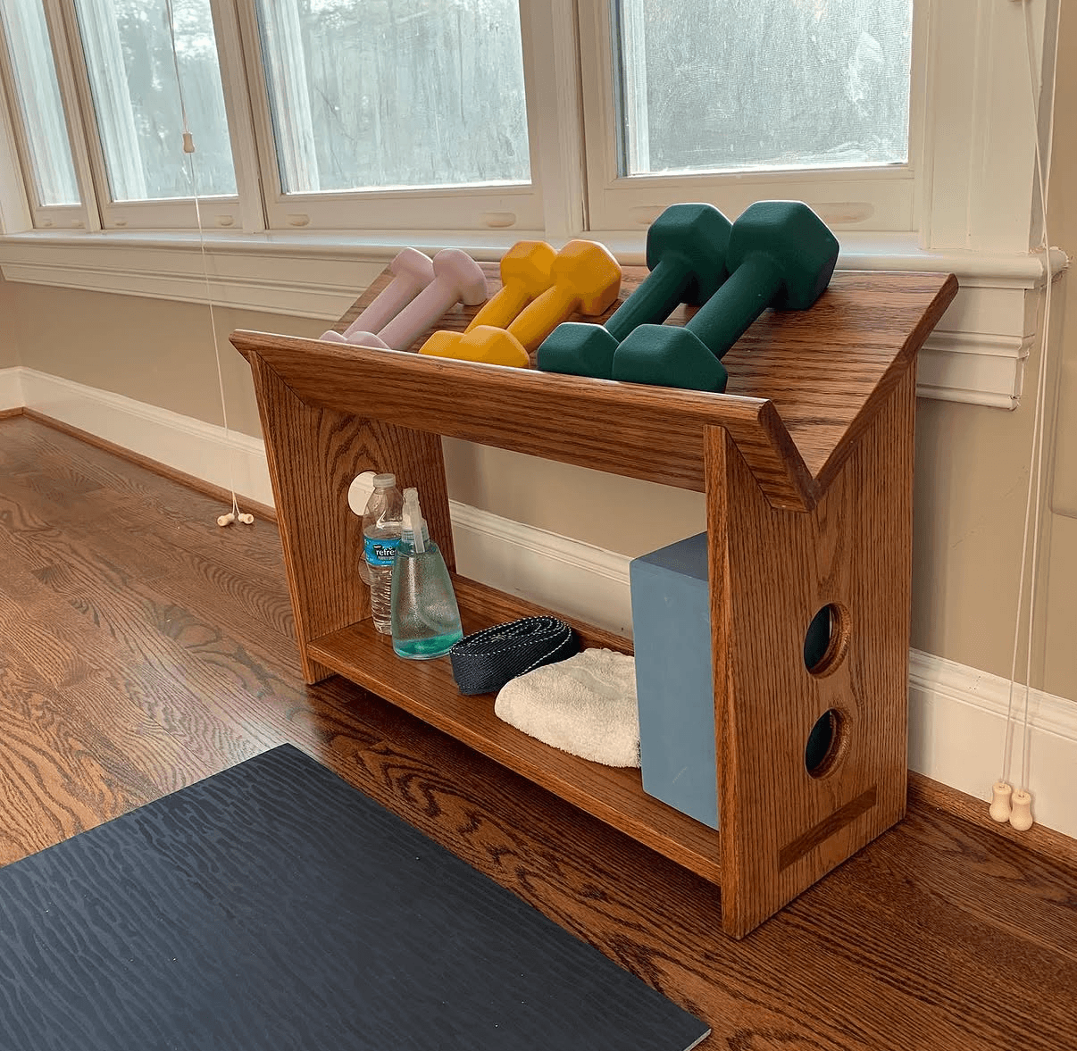 Wooden gym shelf holding hand weights and exercise accessories beneath a window